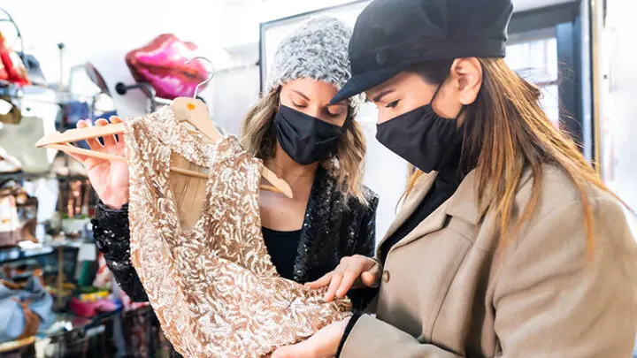 Two shoppers wearing masks inspect clothes in thrift store.