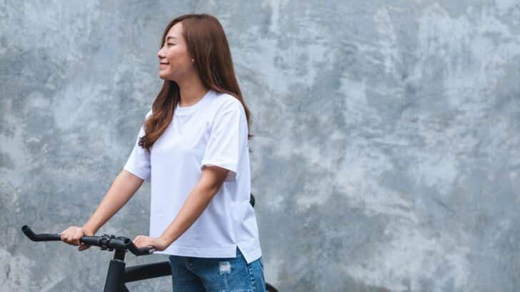 Woman wearing white tee shirt standing outside with bicycle.