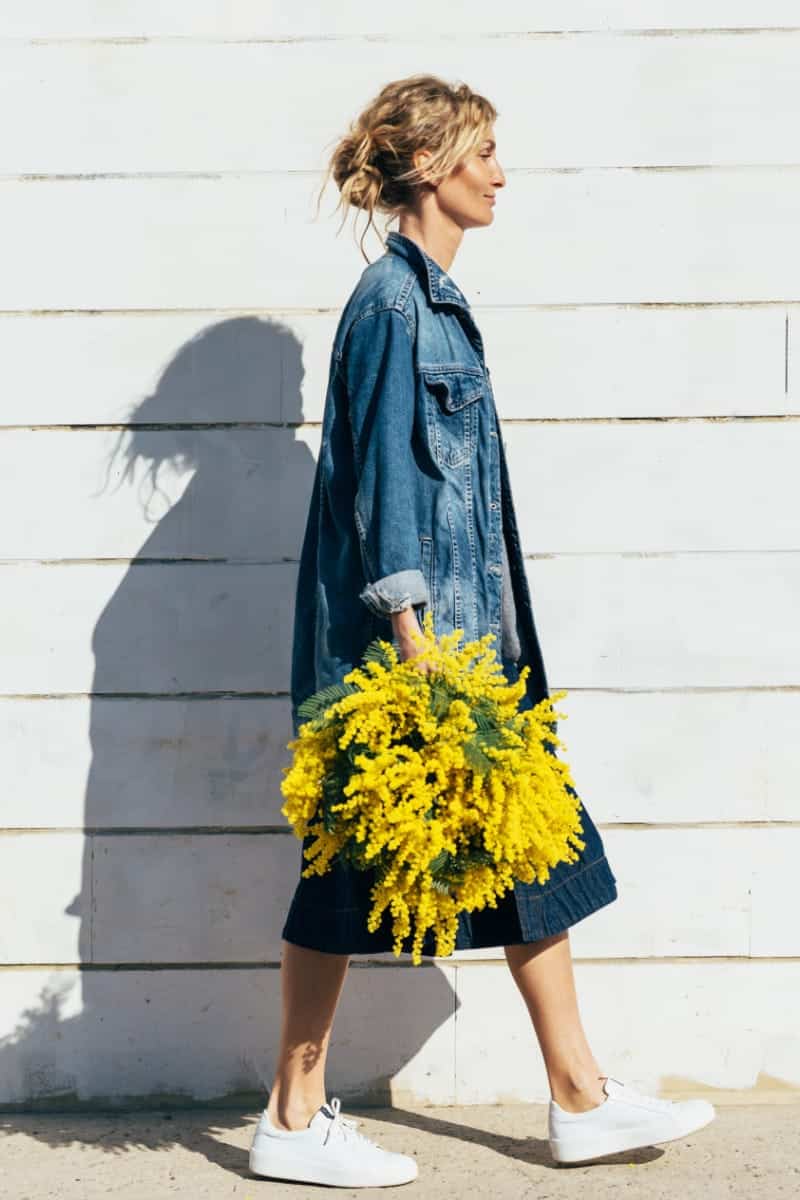 Woman wearing fall denim pieces walks in front of wall holding flowers.