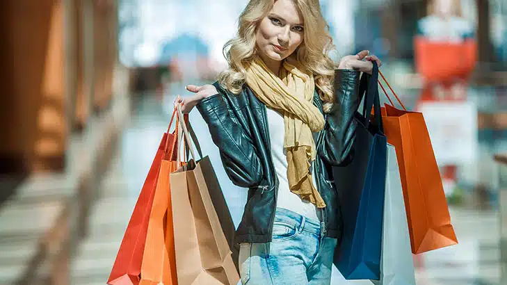 Fashionable woman wearing jeans and scarves holds fashionista gifts in shopping bags outside.