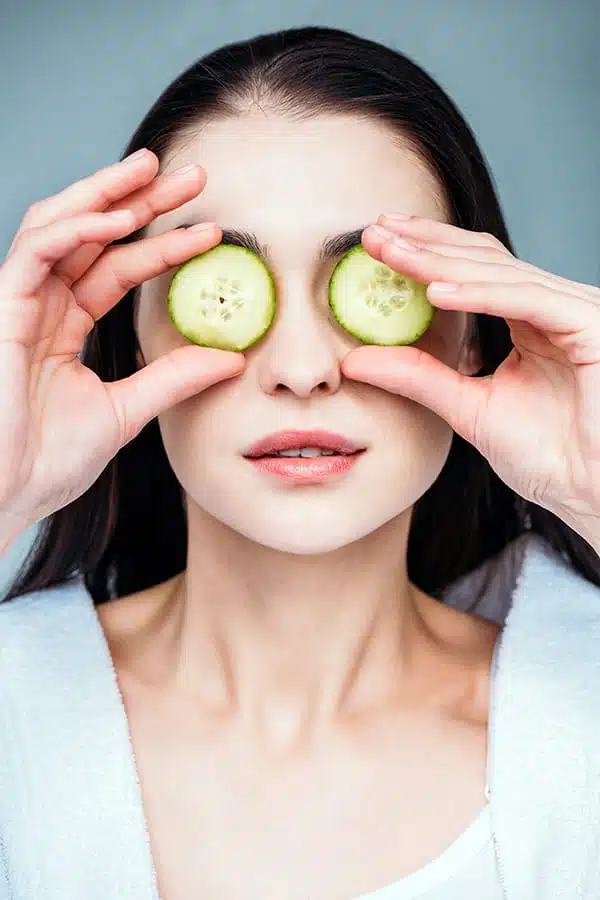 Woman holding cucumbers over her eyes, a skincare home remedy for sagging eyelids.
